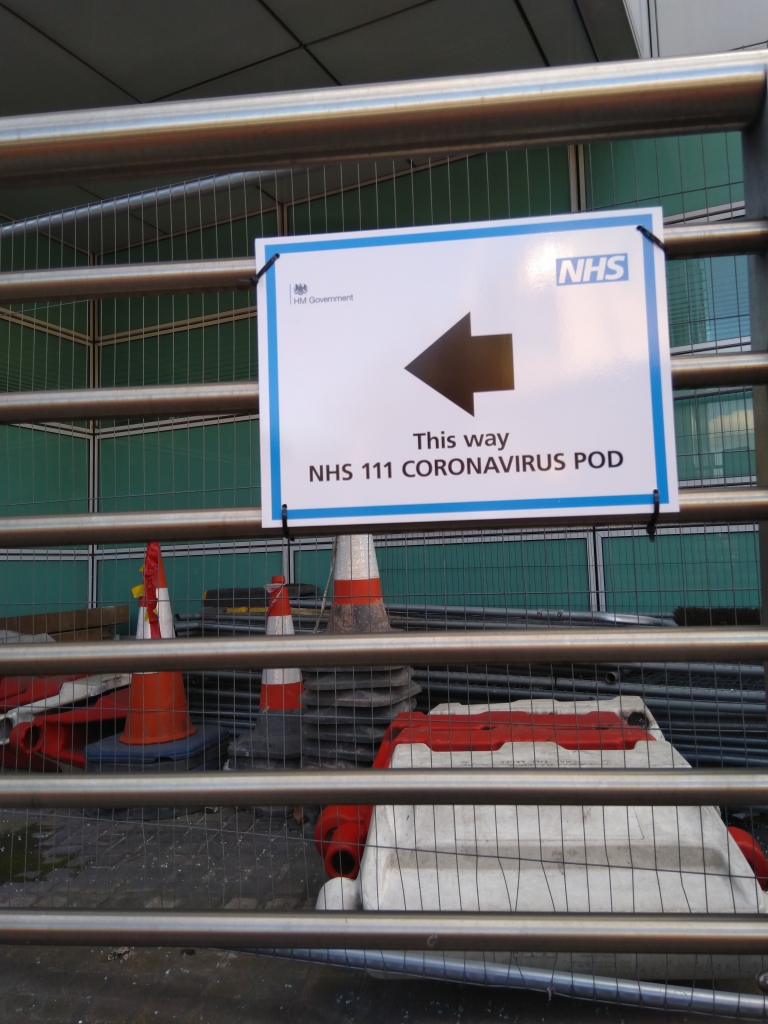 Sign attached by cable ties to railings. The sign reads: "HM Government. NHS. [Left arrow]. This way. NHS 111 CORONAVIRUS POD."